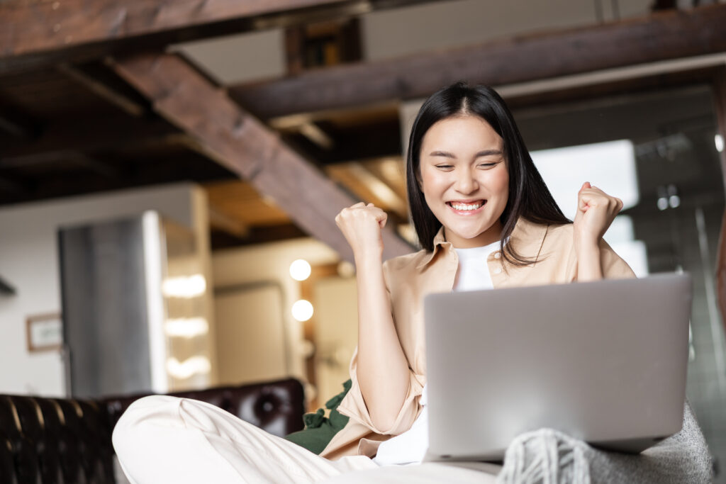 Triumphing asian girl winning online, celebrating victory while watching video on laptop at home